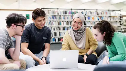 4 students sitting around table in the library looking at laptop.