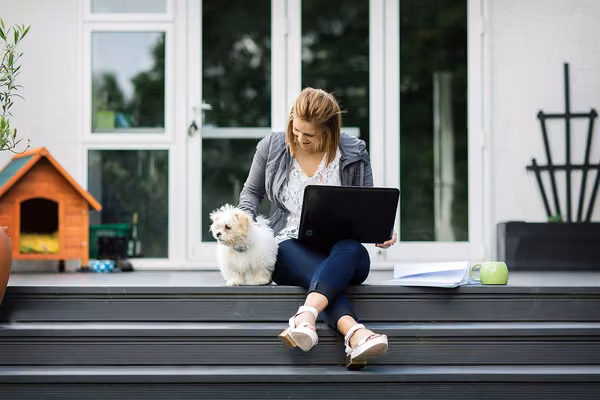 Student sitting on top step, balancing a laptop on their lap, patting a small, white and furry dog