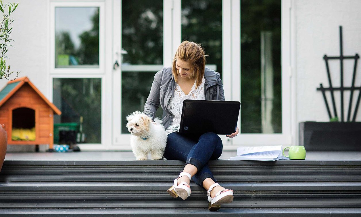 Student sitting on top step, balancing a laptop on their lap, patting a small, white and furry dog