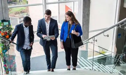 Three people in corporate attire walking up stairs, talking and looking at tablet