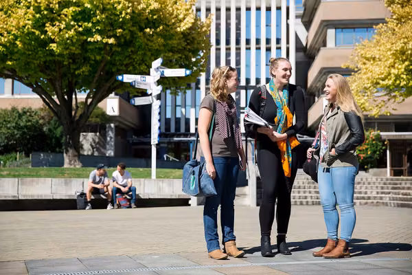 Three students standing on the campus concourse, with buildings and a signpost in the background