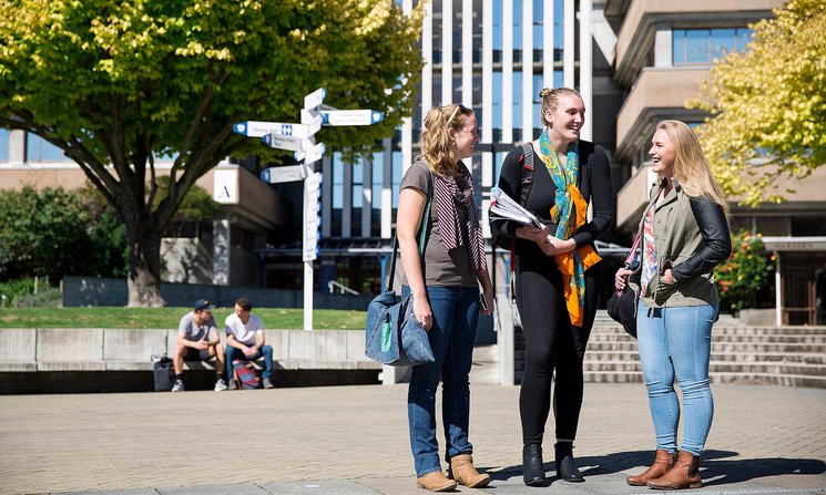 Three students standing on the campus concourse, with buildings and a signpost in the background