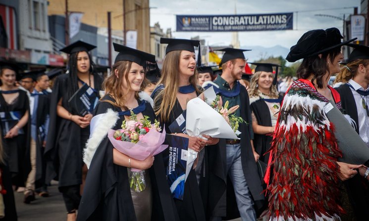Graduates holding large flower bouquets, dressed in graduation attire, walking in a graduation procession
