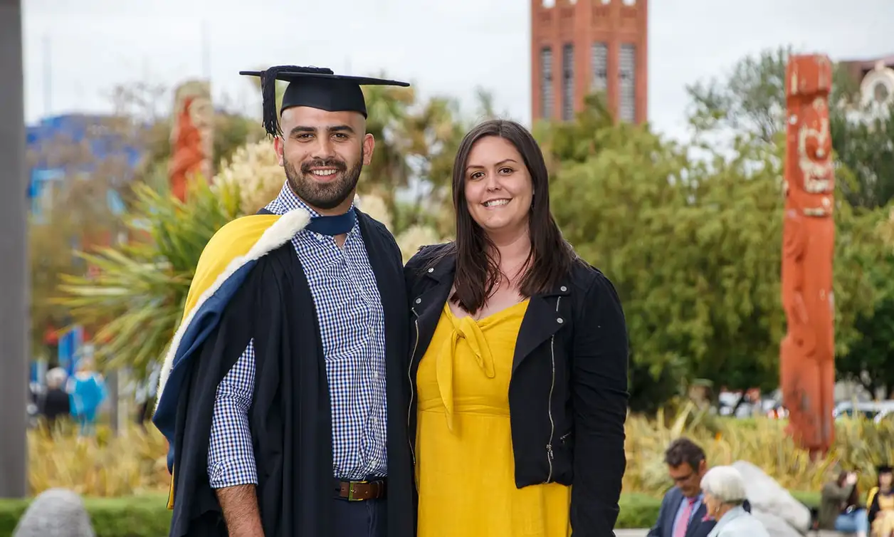 A graduate in graduation attire standing next to a support person, with the campus in the background showing Maori wooden carvings