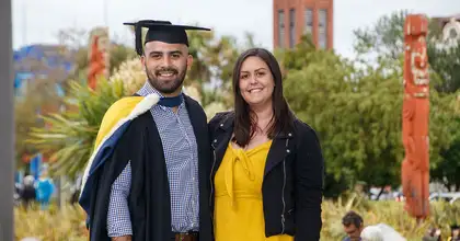 A graduate in graduation attire standing next to a support person, with the campus in the background showing Maori wooden carvings