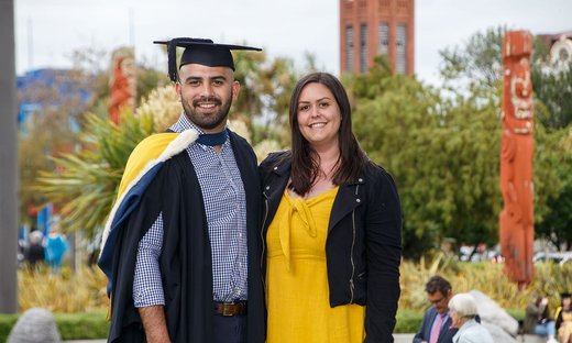 A graduate in graduation attire standing next to a support person, with the campus in the background showing Maori wooden carvings