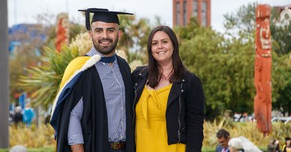 A graduate in graduation attire standing next to a support person, with the campus in the background showing Maori wooden carvings