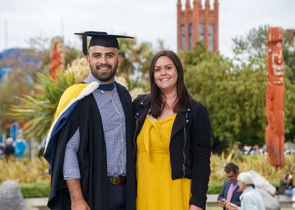A graduate in graduation attire standing next to a support person, with the campus in the background showing Maori wooden carvings