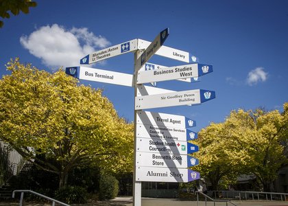 Signpost of building names, with campus buildings and trees in the background