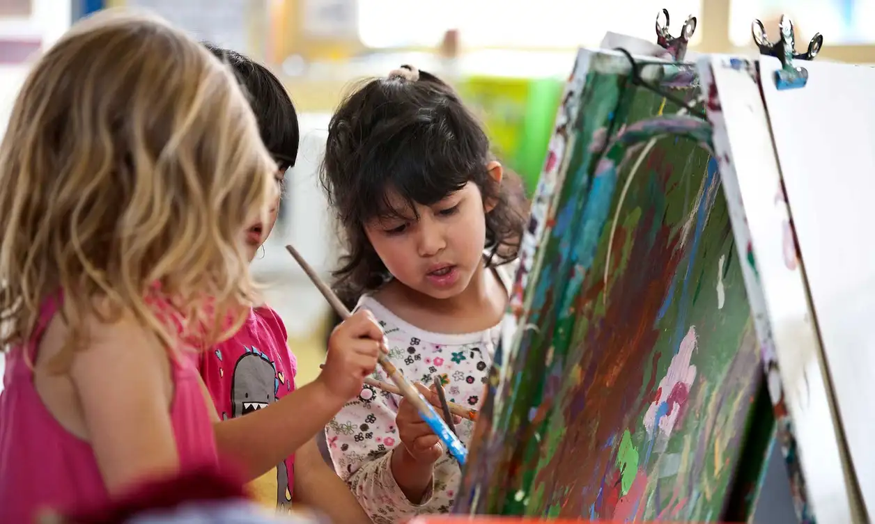 Three children holding paint brushes while painting at Massey University Childcare