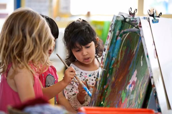 Three children holding paint brushes while painting at Massey University Childcare
