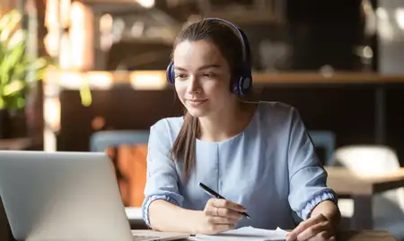 Female student with headphones on studying with a laptop, pen and paper.