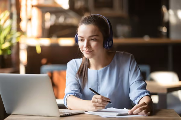 Female student with headphones on studying with a laptop, pen and paper.
