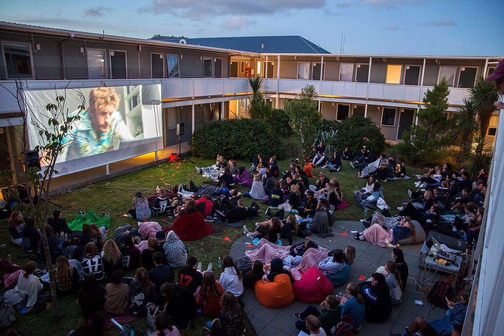 Large gathering of people sitting on the ground watching a large projected screen