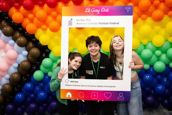 Photo booth at Lil Gay Out featuring three students infront of rainbow balloon display holding an Instagram type frame.