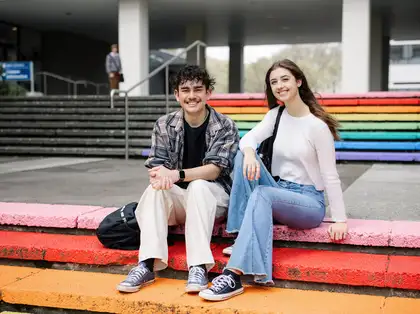 Two students sitting on rainbow painted steps outside Massey University campus.
