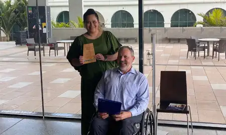 Cerise Tamanui holding her certificate alongside Paul Redmond-Wager who is sitting in a wheelchair.