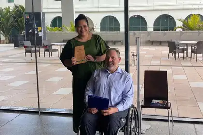 Cerise Tamanui holding her certificate alongside Paul Redmond-Wager who is sitting in a wheelchair.