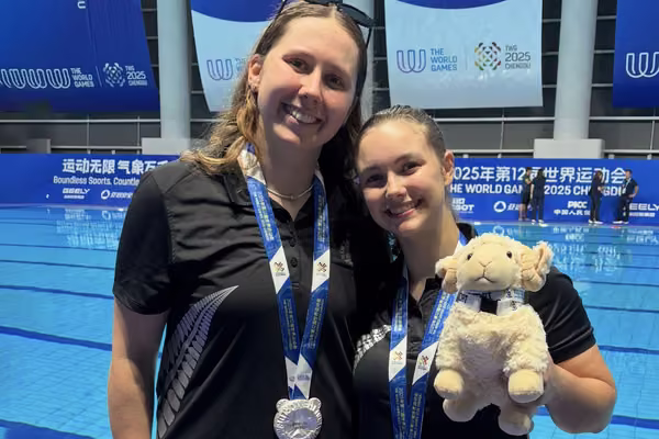 two girls wearing silver medals in front of a pool holding a soft toy ram