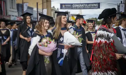 Massey University graduates in academic gowns walking in a street processing, holding flowers.