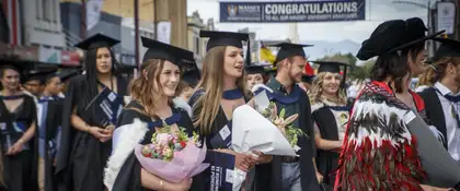 Massey University graduates in academic gowns walking in a street processing, holding flowers.