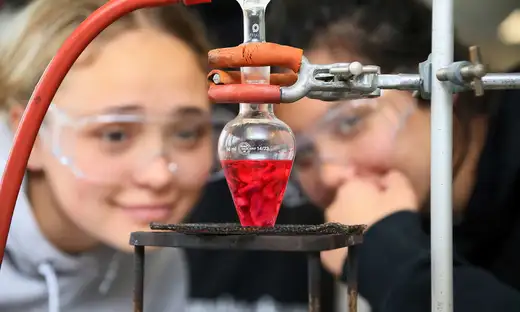 Close-up of two students wearing safety goggles, looking at a science experiment of red liquid in a beaker, over a burner