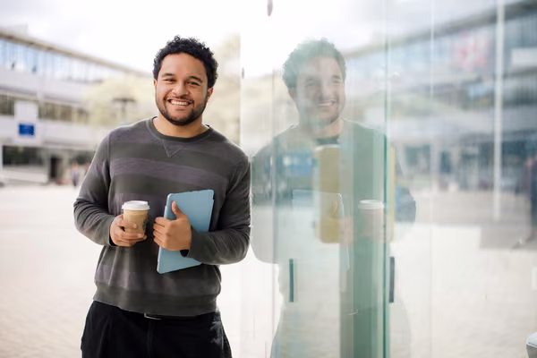 Student standing holding a drink and tablet, leaning on glass with reflection.