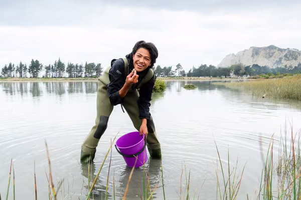 Young person holding a bucket inspects an item from the waterway.