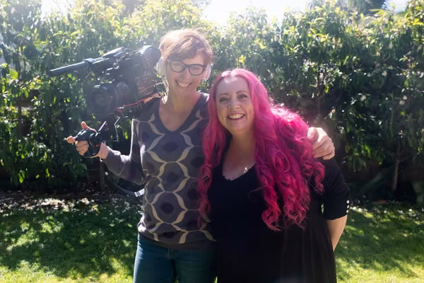 Gwen Isaac holds her camera in one arm, with her other around Siouxsie Wiles as they stand in a garden.