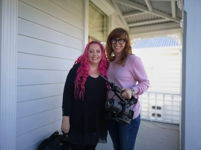 Woman with long pink hair smiling standing with another woman wearing glasses holding a camera on a villa verandah.