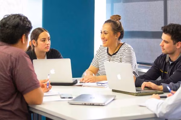 Students sitting in a group around a table talking and smiling with laptops open.