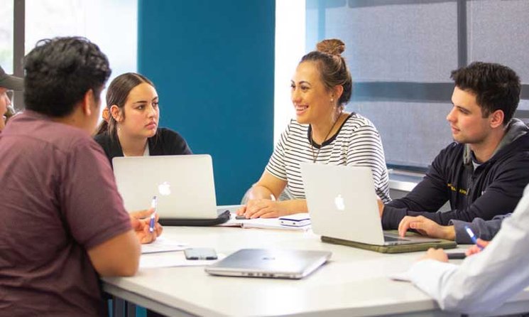 Students sitting in a group around a table talking and smiling with laptops open.
