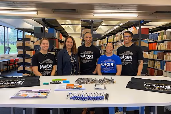 A group of five people from Māori @ �۶���Ƶstand smiling behind a table of stationery