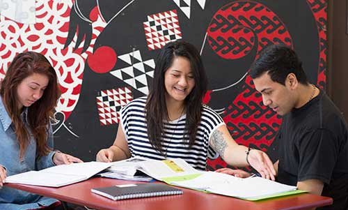 Three students sitting around a table, studying, with a bright red and black wall painting of Maori designs in the background