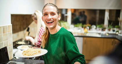Close-up of a person smiling, holding a plate of pasta, inside a kitchen