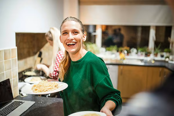 Close-up of a person smiling, holding a plate of pasta, inside a kitchen