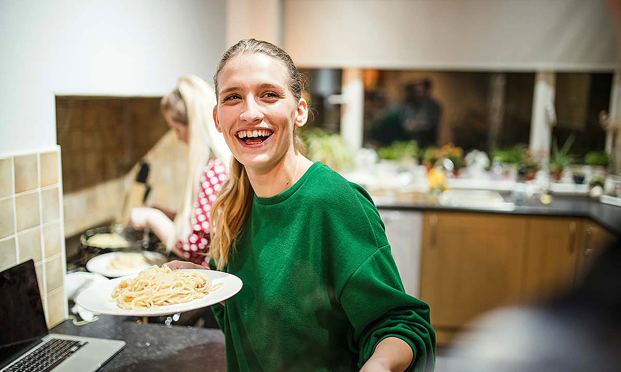 Close-up of a person smiling, holding a plate of pasta, inside a kitchen
