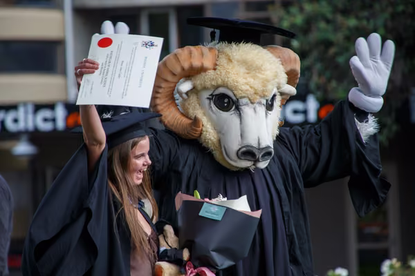 A graduand holds up their graduation certificate while Massey mascot, Fergus, cheers.