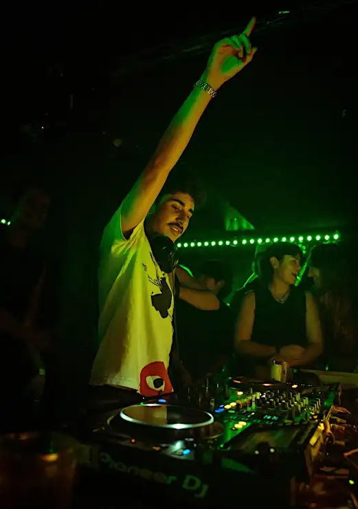 DJ Mikeyy standing behind a turntable in a night club pointing their hand in the air.