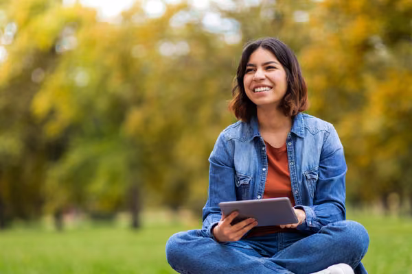 Woman holding tablet sitting in green field.