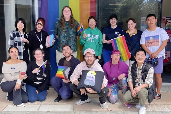 A dozen rainbow advocates standing and crouching in a group. A rainbow flag is behind them.