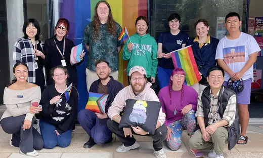 A dozen rainbow advocates standing and crouching in a group. A rainbow flag is behind them.