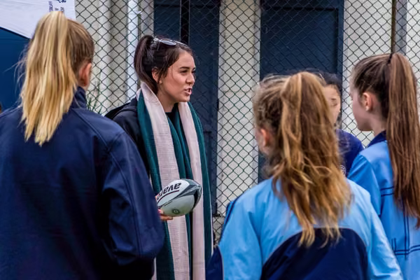 A woman holding a rugby ball speaking to a group of students