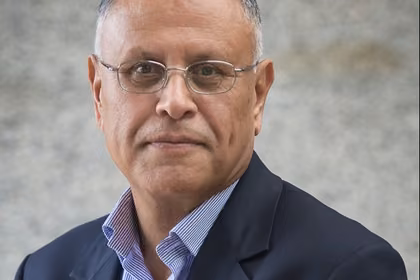 The image shows Professor Fawzi Laswad sitting in front of the camera wearing a dark navy suit.